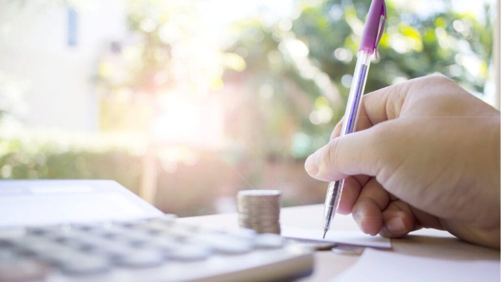 Someone taking notes with a calculator and stack of coins on a table