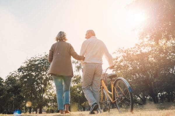 An older couple walking through a park as one pushes a bike